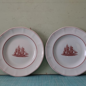 Peut inclure: Deux assiettes en céramique blanche avec un motif de bateau rouge au centre. Les assiettes ont un bord festonné et un bord rouge.