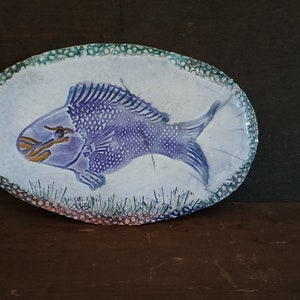 May include: Oval ceramic dish with a hand-painted purple fish design. The fish has detailed scales and is set against a white background with a textured, dotted border in green and pink. The dish is on a dark wooden surface.