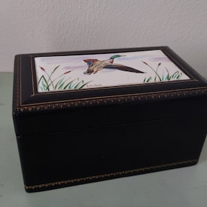 Puede incluir: Una caja de madera negra con una tapa con bisagras. La tapa está decorada con una imagen pintada de un pato silvestre en vuelo sobre un pantano. La caja tiene un borde dorado alrededor de los bordes.
