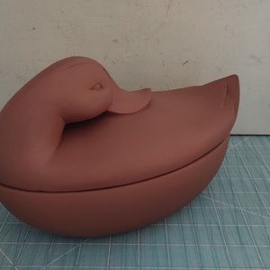 May include: A brown ceramic duck-shaped dish with a lid. The dish is closed and the duck's head is resting on the lid.