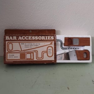 Può includere: Un set di accessori da bar vintage in una scatola di legno. La scatola include un cavatappi, un apribottiglie e una sega per tagliare limoni, lime, arance e formaggio. Il testo "BAR ACCESSORIES" è stampato sulla scatola.