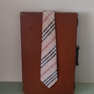 May include: A tan, red, and blue striped tie hangs over a brown, rectangular suitcase. The tie has a classic pattern with diagonal stripes. The suitcase has a handle and a latch. The background is a plain white wall and a green surface.