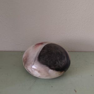 May include: A ceramic decorative stone with a smooth, oval shape. The stone features a dark, textured area contrasted by white and reddish-brown sections. It sits on a light green surface.