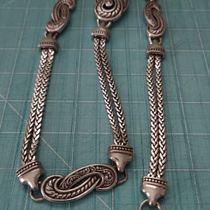 Peut inclure: Un collier en argent avec un pendentif d&eacute;coratif et orn&eacute;. La cha&icirc;ne est compos&eacute;e de deux brins de m&eacute;tal tress&eacute;.