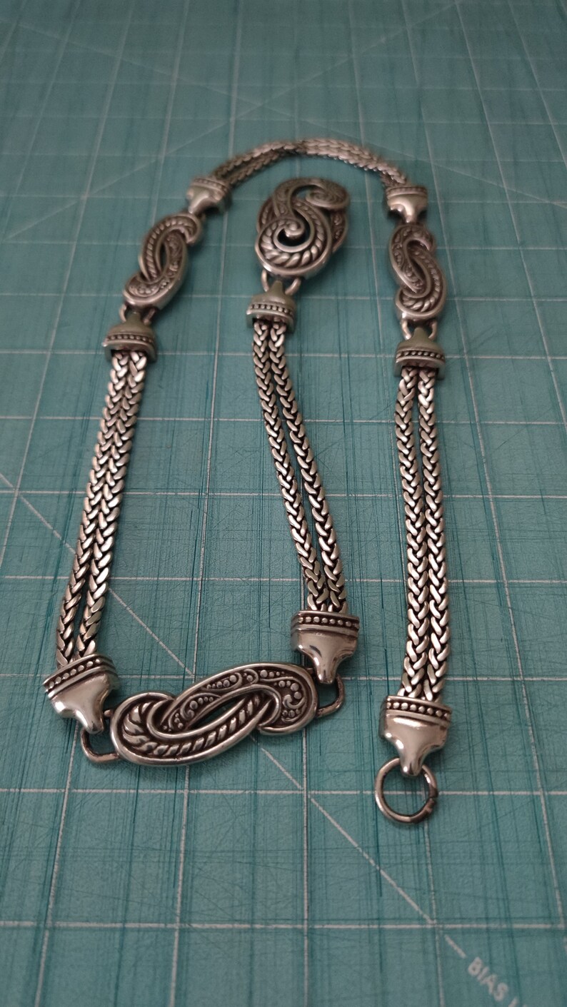 Peut inclure: Un collier en argent avec un pendentif d&eacute;coratif et orn&eacute;. La cha&icirc;ne est compos&eacute;e de deux brins de m&eacute;tal tress&eacute;.