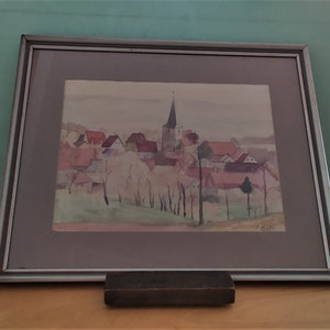 May include: A framed watercolor painting depicting a small village with a church steeple in the center. The houses are painted in shades of pink, red, and brown, and the trees are a soft green. The painting is signed "C. Hefter" in the lower right corner.