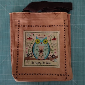 May include: A pink canvas tote bag with brown straps. The bag features a colorful owl with the text "Be Happy. Be Wise." in a white rectangle.