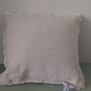 K&ouml;nnte beinhalten: Ein quadratisches, cremefarbenes Kissen mit strukturiertem Stoff und ausgefranstem Rand. Das Kissen liegt auf einer hellgr&uuml;nen Oberfl&auml;che. Das Kissen hat eine neutrale Farbe und w&uuml;rde viele Einrichtungsstile erg&auml;nzen.
