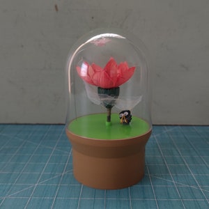 以下が含まれることがあります： ピンクの花がガラスのドームに入っています。緑色の土台に小さなフクロウの置物が置かれています。ドームは青と白の格子状の表面の上にあります。