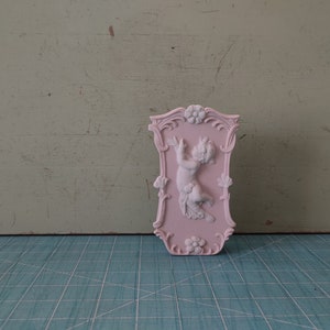 Può includere: Una placca da parete in ceramica rosa con un design in rilievo di un cherubino che tiene un fiore. La placca ha un bordo decorativo con accenti floreali.