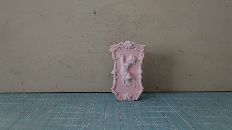 Puede incluir: Una placa de pared de cer&aacute;mica rosa con un dise&ntilde;o en relieve de un querub&iacute;n sosteniendo una flor. La placa tiene un borde decorativo con acentos florales.