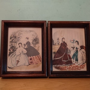 May include: Two framed vintage prints of women in 19th century clothing. The prints are in brown wood frames with a dark brown mount. The print on the left shows two women in a garden setting. The print on the right shows three women in a room setting.