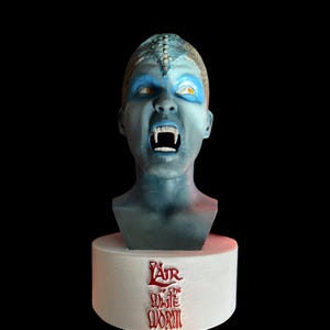 May include: A blue-skinned bust of a creature with sharp fangs and yellow eyes. The bust is mounted on a white cylindrical base with the text "Lair of the White Worm" in red. The background is black.