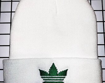 May include: White beanie with a green cannabis leaf logo and the word "cannabis" embroidered below. The beanie is made of a soft, knitted material and has a folded brim. The logo is a stylized cannabis leaf.