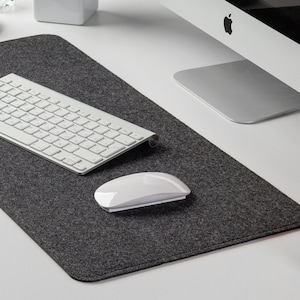 May include: A gray felt desk mat with a white keyboard and a white wireless mouse. The mat is on a white desk with a computer monitor, a potted plant, and a decorative lamp.