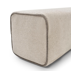 Puede incluir: Primer plano de un cojín rectangular de tela beige con una superficie texturizada. Los bordes están delineados con un ribete marrón-gris oscuro. El cojín tiene esquinas redondeadas y un diseño minimalista.