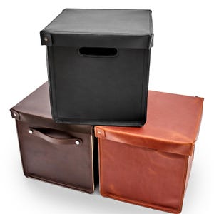 Peut inclure: Trois boîtes de rangement en cuir noir, marron et brun rougeâtre. Chaque boîte a un couvercle, une poignée et est de forme carrée. Les boîtes sont empilées, la boîte noire étant au-dessus. Les poignées sont fixées avec des rivets argentés.