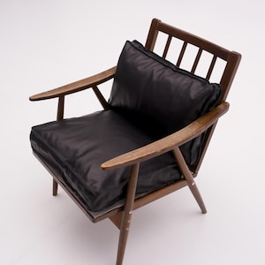 Puede incluir: Un sillón moderno de mediados de siglo con estructura y reposabrazos de madera oscura. La silla tiene un cojín y respaldo negros. La silla está sobre un fondo blanco.