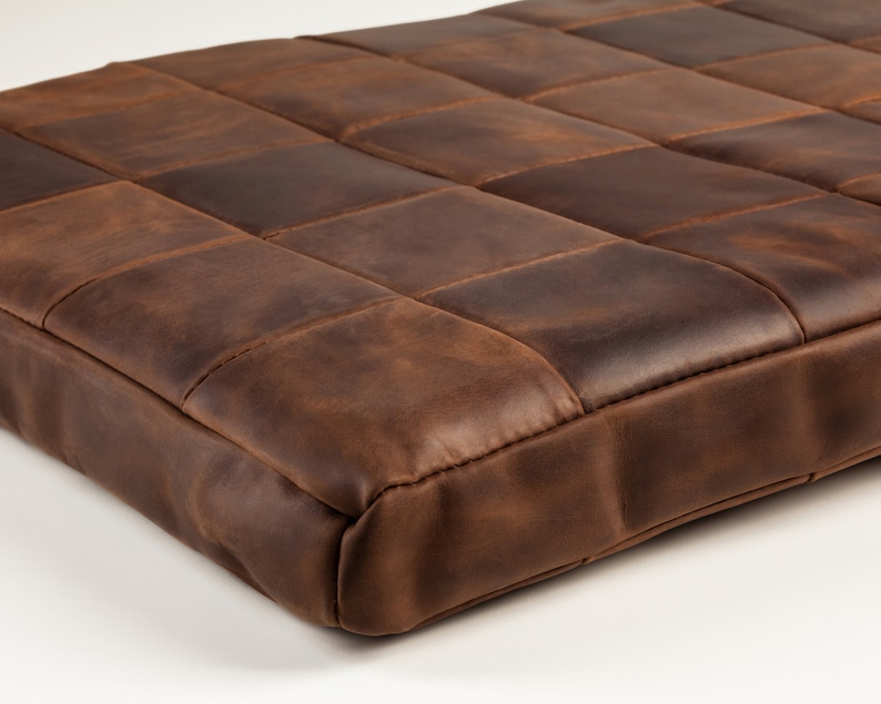 Op de afbeelding: Een vierkant, bruin leren kussen met een patchwork-ontwerp. Het kussen is verdeeld in rechthoekige secties, elk met een iets andere bruintint, waardoor een textuur ontstaat. De randen zijn netjes gestikt en de algehele esthetiek is rustiek en duurzaam.