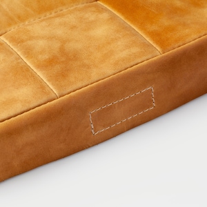 May include: Close-up of a section of a tan leather cushion. The cushion is divided into rectangular sections with stitched seams. The edges are a darker shade of tan, and a rectangular tag is stitched on the side.
