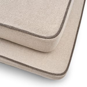 Könnte beinhalten: Nahaufnahme von zwei beigefarbenen Kissen mit strukturierter Oberfläche und dunkelbraunem Rand. Die Kissen sind gestapelt, wobei eines teilweise unter dem anderen sichtbar ist. Der Stoff wirkt weich und einladend.