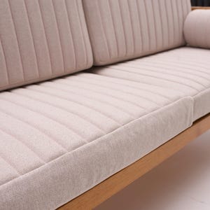 May include: A light beige sofa with a wooden frame. The sofa features a tufted design on the seat and back cushions, and a cylindrical bolster pillow. The wooden frame has a natural finish.