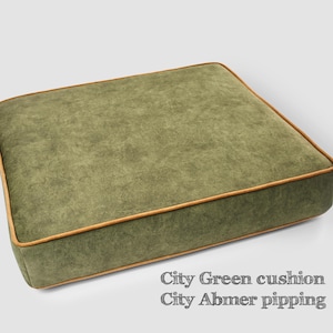 May include: A square, olive green cushion with a textured surface and a light brown trim. The cushion has the text "City Green cushion City Abmer pipping" in the bottom right corner.