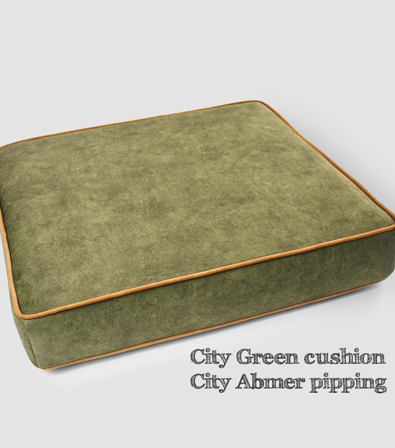 May include: A square, olive green cushion with a textured surface and a light brown trim. The cushion has the text "City Green cushion City Abmer pipping" in the bottom right corner.