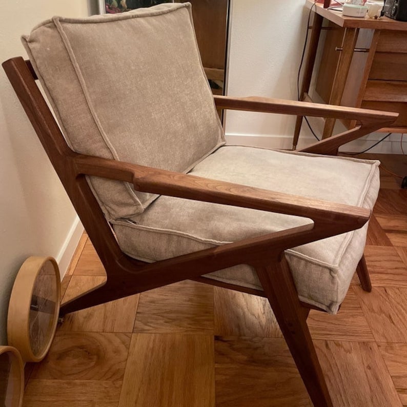 May include: A mid-century modern armchair with a light beige cushion and backrest. The chair features a dark brown wooden frame with angled armrests and legs. The design is minimalist and stylish.