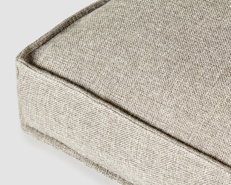 May include: Close-up of a square, light beige cushion with a textured, woven fabric. The edges are neatly sewn with a darker thread, creating a defined border. The neutral color and simple design suggest a focus on comfort and versatility.