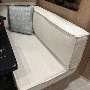 May include: A built-in bench seat with a light beige fabric covering the seat and backrest. A square, light blue patterned pillow rests against the backrest. The bench is part of a seating area, possibly in a recreational vehicle.
