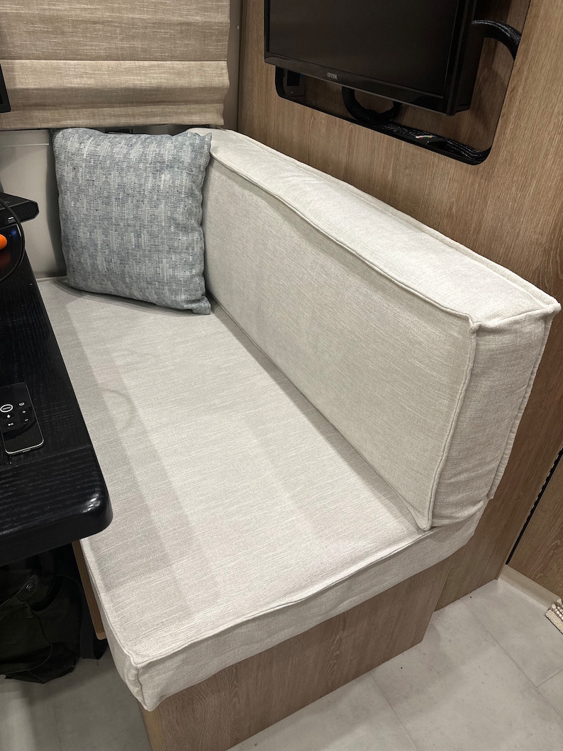 May include: A built-in bench seat with a light beige fabric covering the seat and backrest. A square, light blue patterned pillow rests against the backrest. The bench is part of a seating area, possibly in a recreational vehicle.
