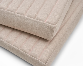 May include: Two beige square cushions with vertical, pale pink stripes. The cushions have a thick, textured fabric and a neat, tailored appearance. The cushions are stacked, highlighting their design and construction.