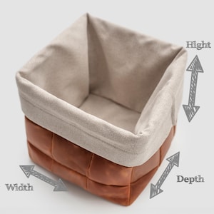 May include: A square storage basket with a brown leather exterior and a beige fabric interior. The leather is stitched in a grid pattern. The basket has a folded-over top edge. The image shows the height, width, and depth of the basket.