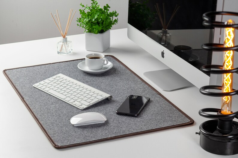 May include: A gray felt desk mat with brown leather trim, a white wireless keyboard, a white wireless mouse, a black smartphone, and a white coffee cup on a white desk.