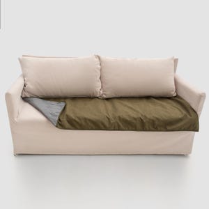 Può includere: Un divano beige chiaro con due cuscini rettangolari abbinati e una coperta verde oliva scuro drappeggiata sul sedile. La coperta ha un lato inferiore grigio. Il divano è su uno sfondo bianco.