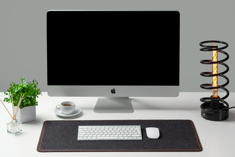 May include: A white computer monitor with a black screen, a white keyboard, a white mouse, a white coffee cup and saucer, a potted green plant, and a black lamp with a coiled metal base and a clear light bulb on a white desk.