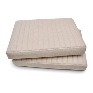 Puede incluir: Dos cojines cuadrados beige apilados uno encima del otro. Los cojines tienen un patrón de rayas verticales acolchadas. Los bordes de los cojines son de color beige liso. El fondo es blanco.
