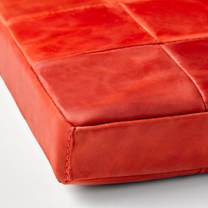Op de afbeelding: Close-up van een levendig rood, vierkant kussen. Het oppervlak is verdeeld in gestikte vierkanten, met naden zichtbaar langs de randen. Het materiaal lijkt leer of een vergelijkbare textuurstof te zijn.