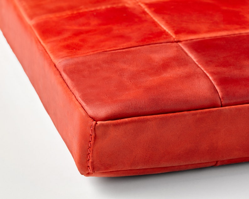 Op de afbeelding: Close-up van een levendig rood, vierkant kussen. Het oppervlak is verdeeld in gestikte vierkanten, met naden zichtbaar langs de randen. Het materiaal lijkt leer of een vergelijkbare textuurstof te zijn.