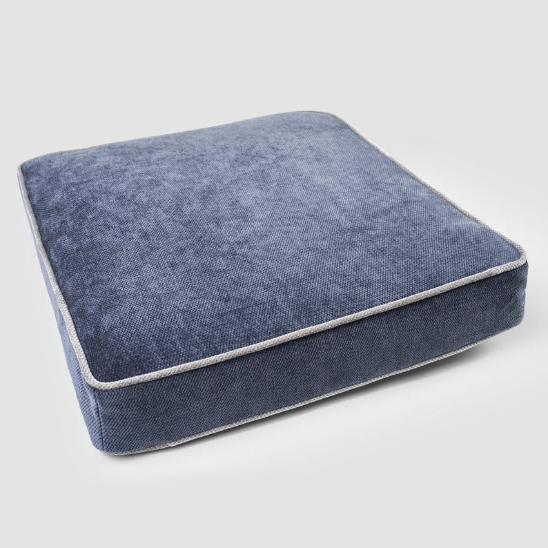 May include: Square blue cushion with a textured fabric and a light gray trim. The cushion is a solid color and appears to be made of a soft material. The cushion is designed for seating.