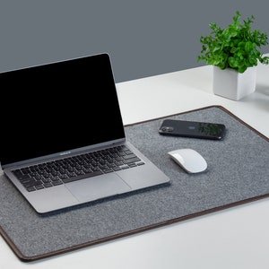 May include: A gray felt desk mat with brown leather trim. A laptop, smartphone, and wireless mouse are placed on the mat.