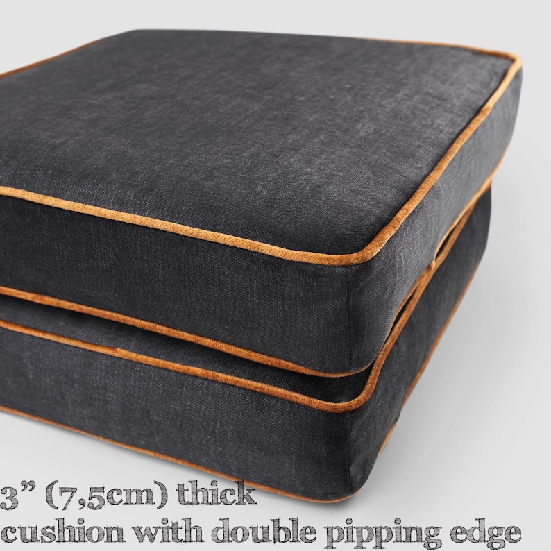 May include: Two square, charcoal-colored cushions with a 3-inch thickness and double piping edges. The edges are trimmed with a contrasting gold-colored piping. The cushions are stacked on top of each other.