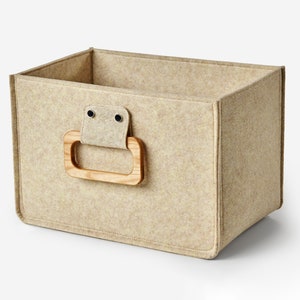Könnte beinhalten: Ein beigefarbener Aufbewahrungskorb aus Filz mit einem Holzgriff. Der Korb hat ein schlichtes Design und ist perfekt zum Organisieren kleiner Gegenstände.