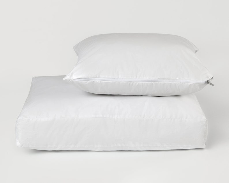 May include: A white zippered pillow protector sits on top of a white zippered mattress protector. Both are made of a soft, smooth fabric.