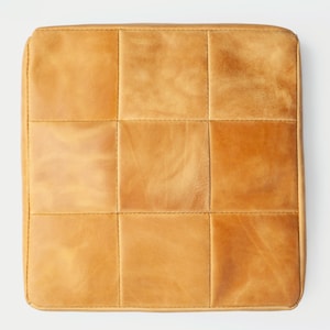 May include: Square, tan leather cushion with a grid pattern created by stitched seams. The cushion has a slightly worn appearance, with variations in color and texture across its surface. The seams are a lighter tan color.