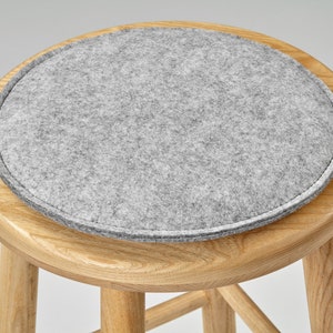 May include: A grey felt cushion sits atop a wooden stool. The stool has a round seat and four legs.