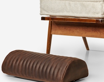 May include: A brown leather footrest with a quilted design. The footrest is in front of a wooden chair with a white cushion.