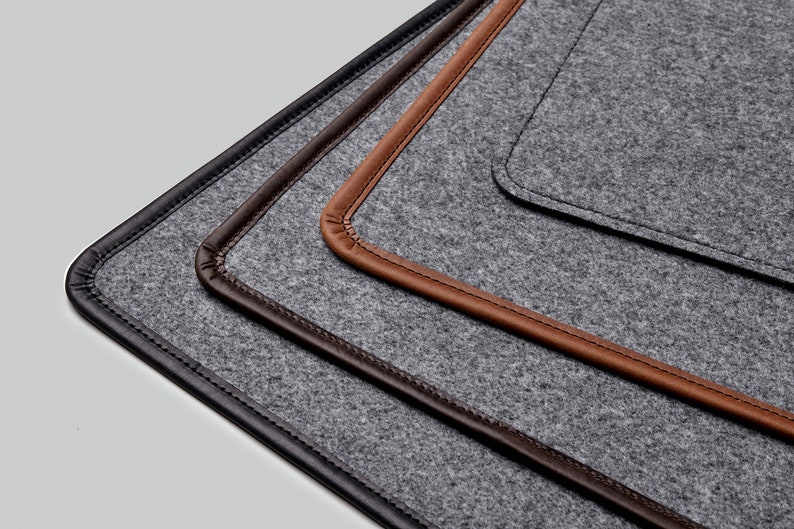 May include: Three gray felt placemats with different colored leather trim. The placemats are stacked on top of each other, with the black leather trim on the bottom, the brown leather trim in the middle, and the tan leather trim on top.