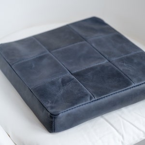 May include: A square, navy blue cushion with a quilted design. The cushion is made of a leather-like material and has a stitched border. The cushion is placed on a white surface.